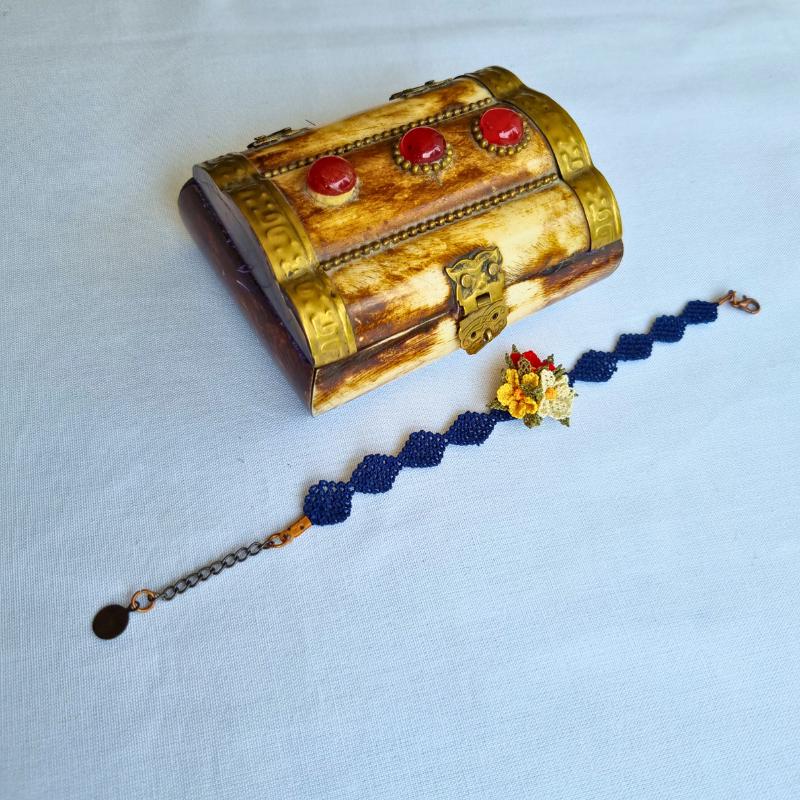 A handmade bracelet with three needle lace flowers in red, ivory, and yellow, with green leaves, on a dark blue band. Made with artificial silk thread using the Turkish Oya technique.