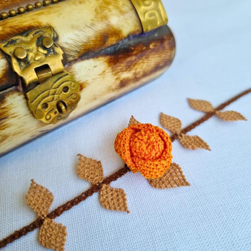 A handmade bracelet featuring a single orange rose with brown needle lace leaves on a brown band, made with artificial silk thread using the traditional Turkish Oya technique.