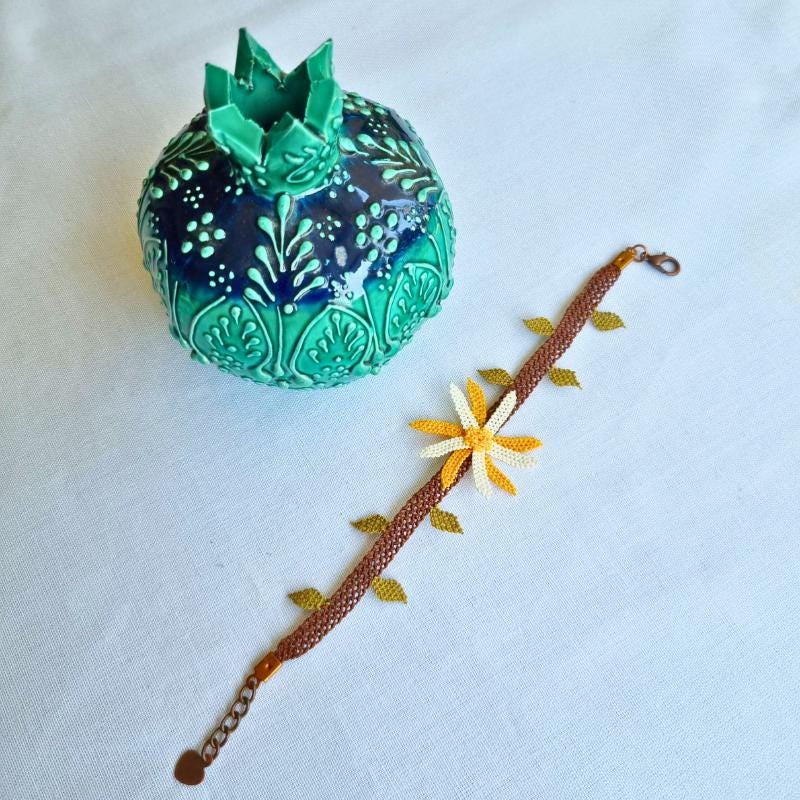 A handmade bracelet featuring one yellow and ivory needle lace flower with green leaves on a brown band. Made from artificial silk thread in the traditional Turkish Oya style.