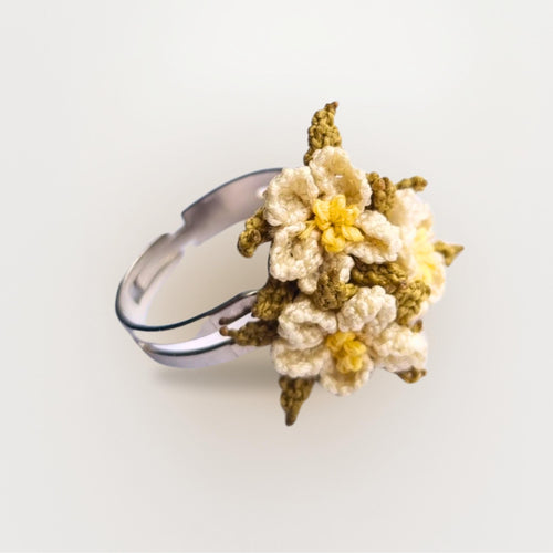 handmade adjustable ring with cream needle lace flowers, yellow centers, and green leaf details. Made from artificial silk thread. Worn on a hand.