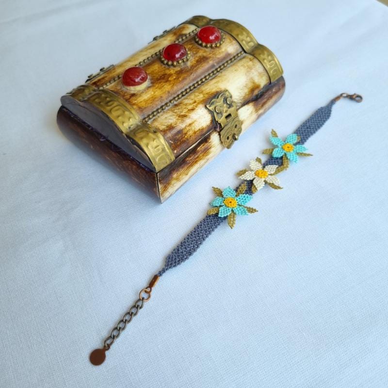 A handmade bracelet featuring three turquoise blue needle lace flowers with golden yellow centers, ivory accents, and olive green leaves, arranged on a lavender purple adjustable band. Made using artificial silk thread in the traditional Turkish Oya style.