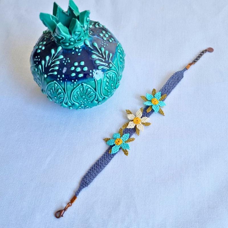 A handmade bracelet featuring three turquoise blue needle lace flowers with golden yellow centers, ivory accents, and olive green leaves, arranged on a lavender purple adjustable band. Made using artificial silk thread in the traditional Turkish Oya style.