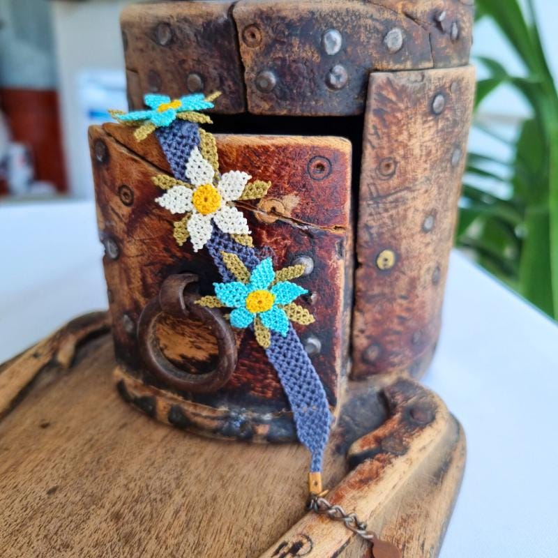 A handmade bracelet featuring three turquoise blue needle lace flowers with golden yellow centers, ivory accents, and olive green leaves, arranged on a lavender purple adjustable band. Made using artificial silk thread in the traditional Turkish Oya style.