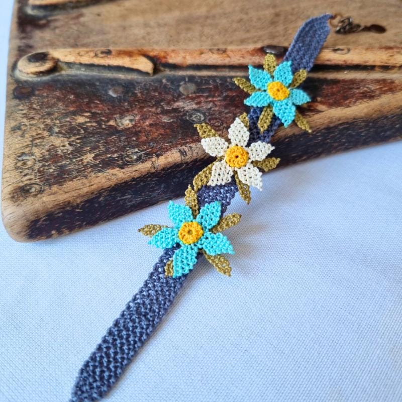 A handmade bracelet featuring three turquoise blue needle lace flowers with golden yellow centers, ivory accents, and olive green leaves, arranged on a lavender purple adjustable band. Made using artificial silk thread in the traditional Turkish Oya style.