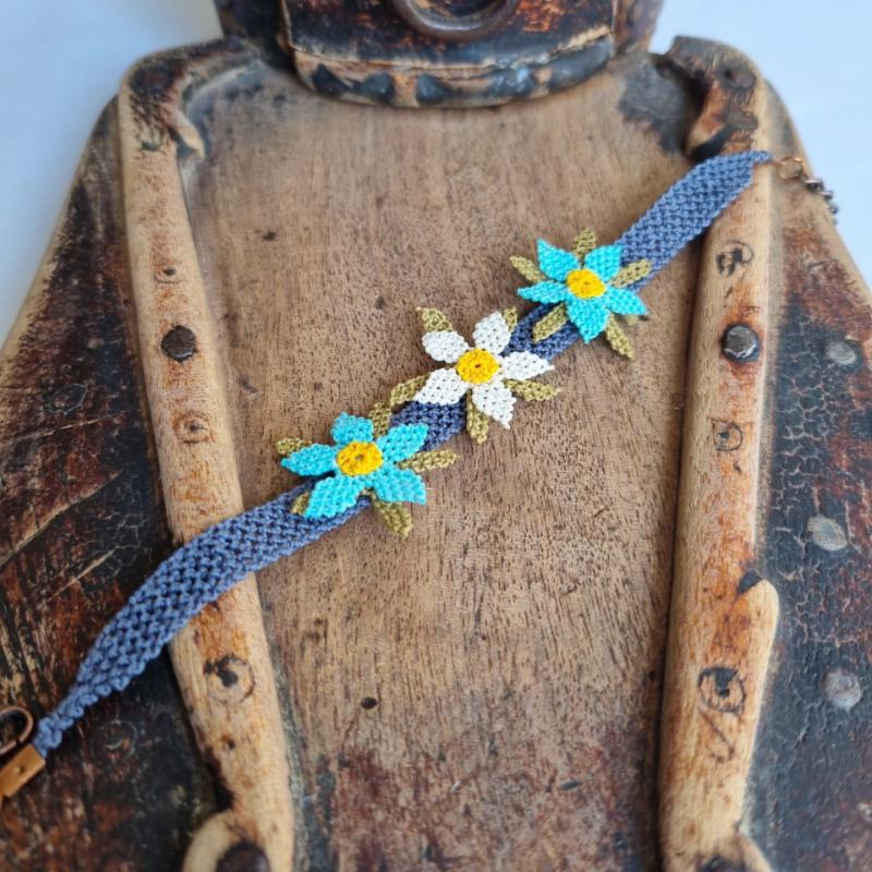 A handmade bracelet featuring three turquoise blue needle lace flowers with golden yellow centers, ivory accents, and olive green leaves, arranged on a lavender purple adjustable band. Made using artificial silk thread in the traditional Turkish Oya style.