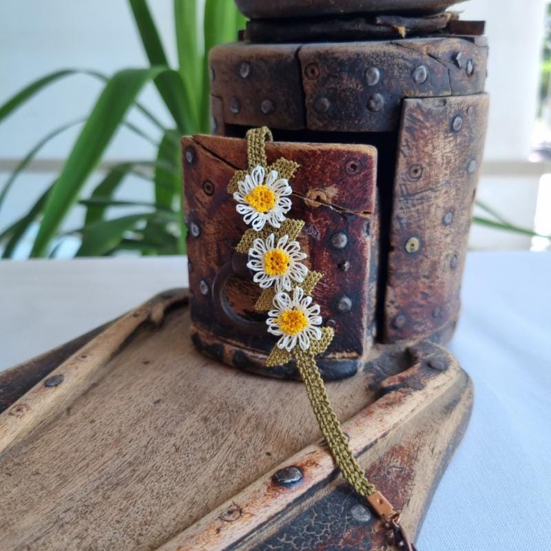 A handmade bracelet with three white daisy flowers made from needle lace, featuring yellow centers and green leaves, set on an olive green adjustable band. Made using artificial silk thread with traditional Turkish Oya technique.