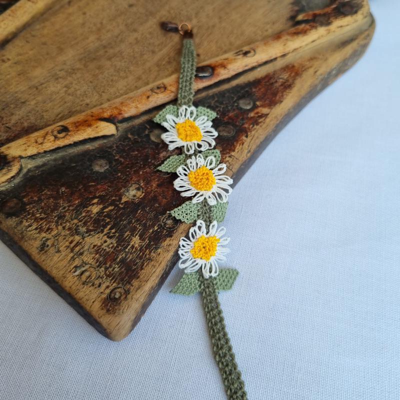 A handmade bracelet with three white daisy flowers made from Turkish needle lace, featuring yellow centers and green leaves, on a light olive green crocheted band. Made from artificial silk thread.