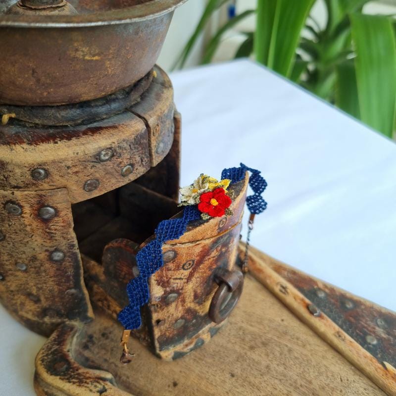 A handmade bracelet with three needle lace flowers in red, ivory, and yellow, with green leaves, on a dark blue band. Made with artificial silk thread using the Turkish Oya technique.