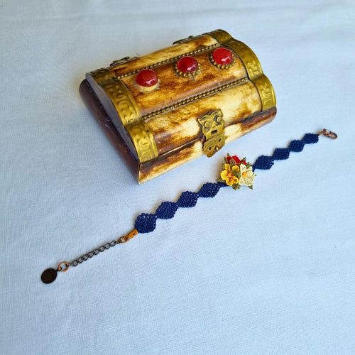 A handmade bracelet with three needle lace flowers in red, ivory, and yellow, with green leaves, on a dark blue band. Made with artificial silk thread using the Turkish Oya technique.