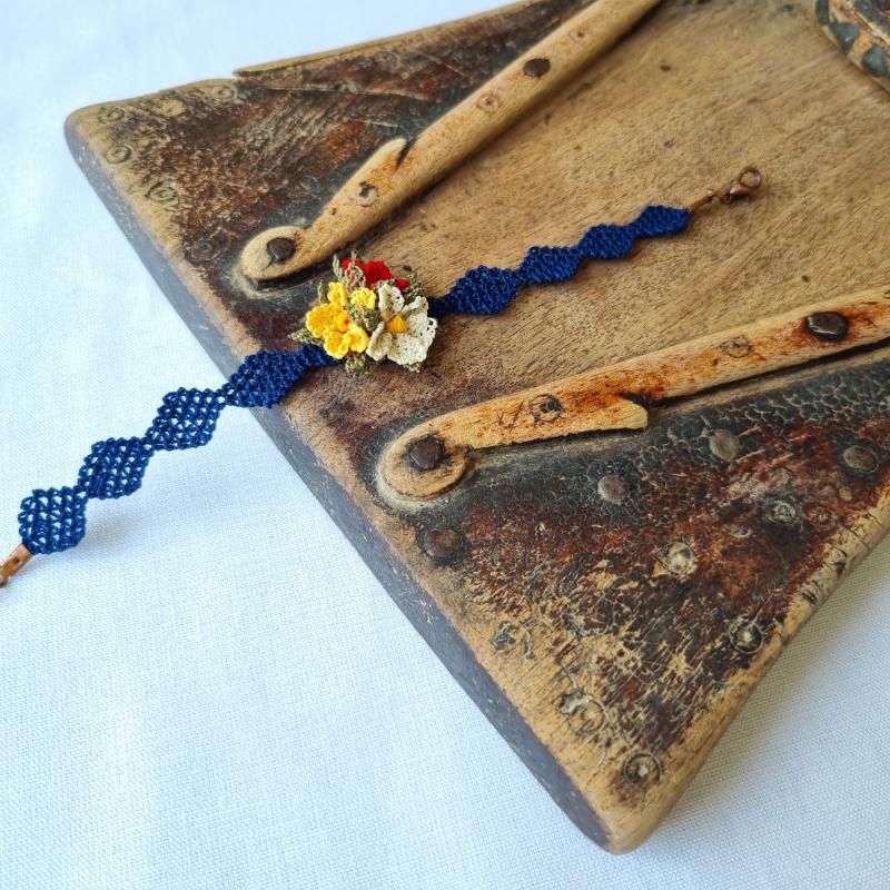 A handmade bracelet with three needle lace flowers in red, ivory, and yellow, with green leaves, on a dark blue band. Made with artificial silk thread using the Turkish Oya technique.