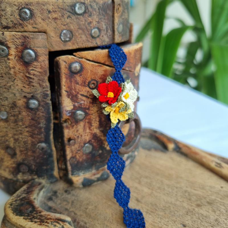 A handmade bracelet with three needle lace flowers in red, ivory, and yellow, with green leaves, on a dark blue band. Made with artificial silk thread using the Turkish Oya technique.