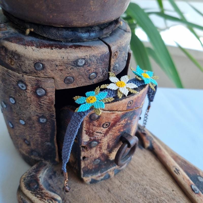 A handmade bracelet featuring three turquoise blue needle lace flowers with golden yellow centers, ivory accents, and olive green leaves, arranged on a lavender purple adjustable band. Made using artificial silk thread in the traditional Turkish Oya style.