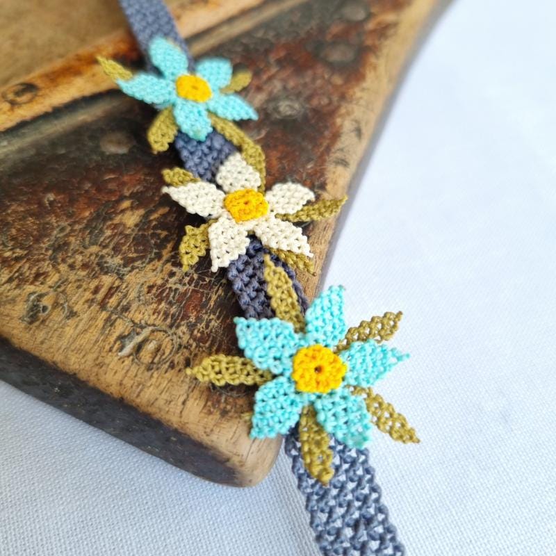 A handmade bracelet featuring three turquoise blue needle lace flowers with golden yellow centers, ivory accents, and olive green leaves, arranged on a lavender purple adjustable band. Made using artificial silk thread in the traditional Turkish Oya style.