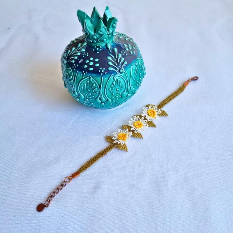 A handmade bracelet with three white daisy flowers made from needle lace, featuring yellow centers and green leaves, set on an olive green adjustable band. Made using artificial silk thread with traditional Turkish Oya technique.