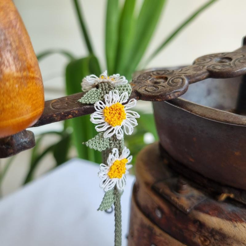 A handmade bracelet with three white daisy flowers made from Turkish needle lace, featuring yellow centers and green leaves, on a light olive green crocheted band. Made from artificial silk thread.