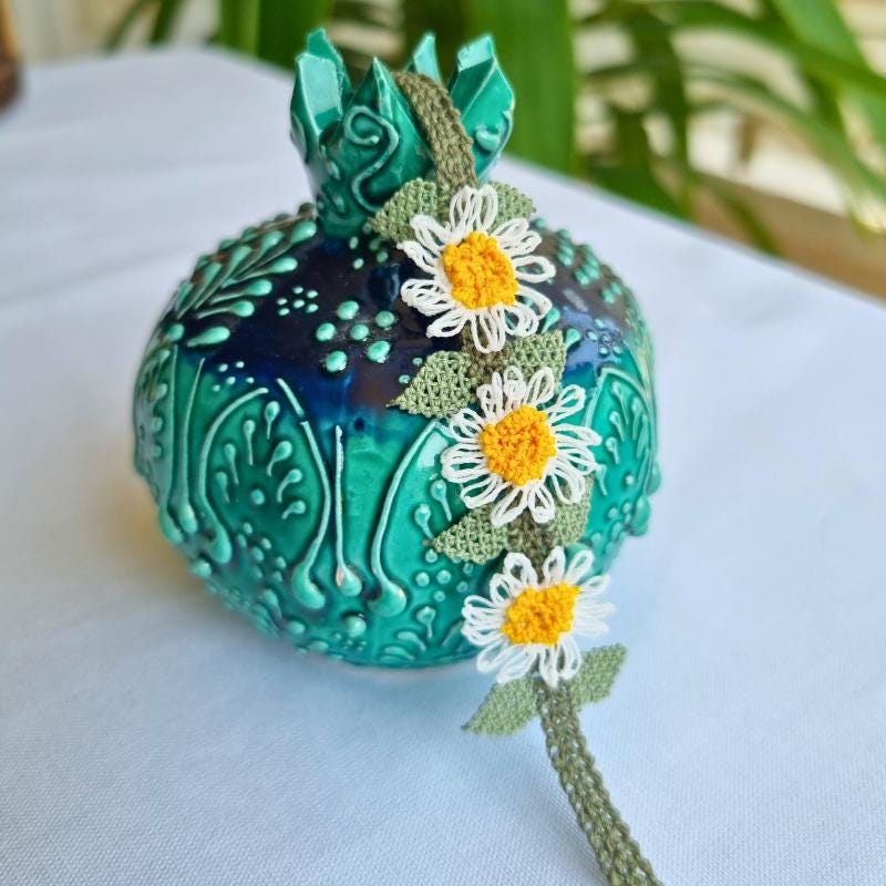 A handmade bracelet with three white daisy flowers made from Turkish needle lace, featuring yellow centers and green leaves, on a light olive green crocheted band. Made from artificial silk thread.