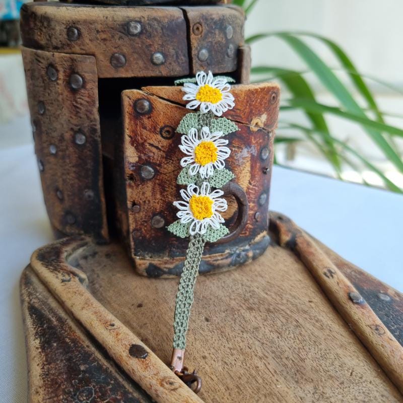 A handmade bracelet with three white daisy flowers made from Turkish needle lace, featuring yellow centers and green leaves, on a light olive green crocheted band. Made from artificial silk thread.