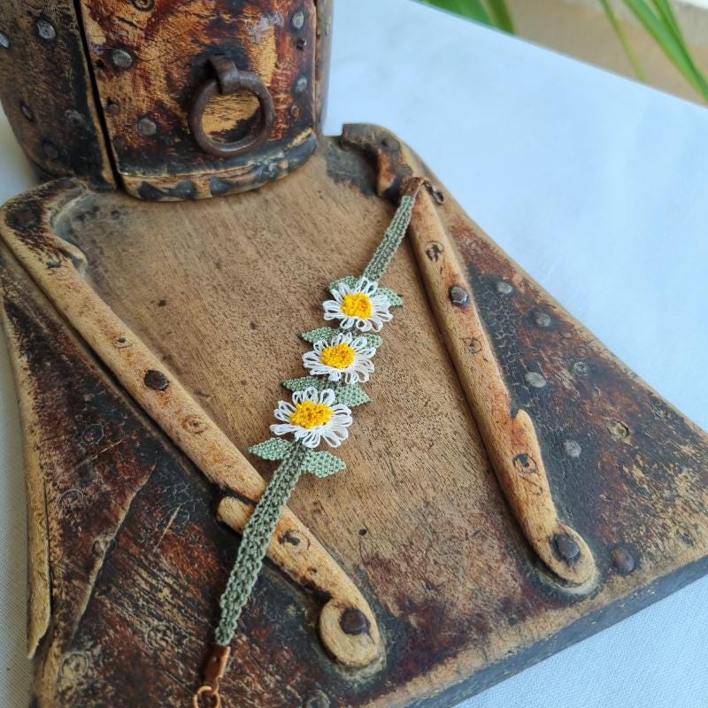 A handmade bracelet with three white daisy flowers made from Turkish needle lace, featuring yellow centers and green leaves, on a light olive green crocheted band. Made from artificial silk thread.