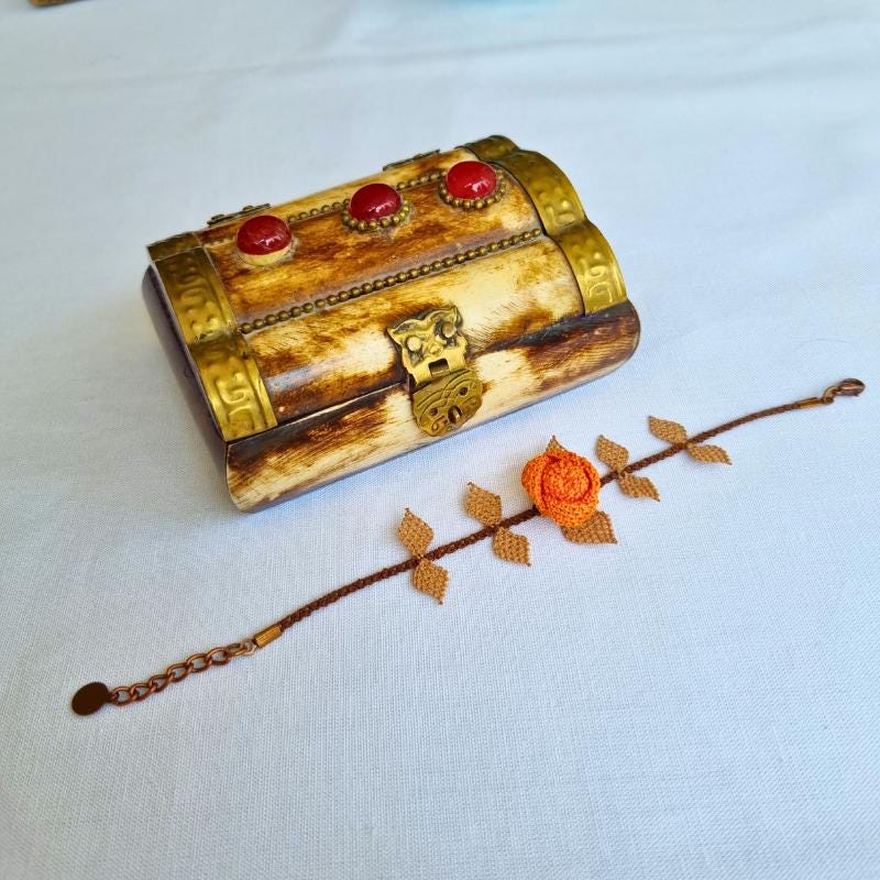 A handmade bracelet featuring a single orange rose with brown needle lace leaves on a brown band, made with artificial silk thread using the traditional Turkish Oya technique.