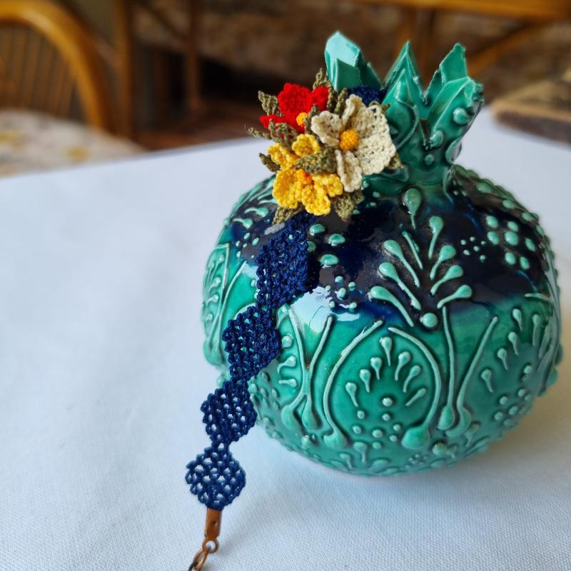 A handmade bracelet with three needle lace flowers in red, ivory, and yellow, with green leaves, on a dark blue band. Made with artificial silk thread using the Turkish Oya technique.