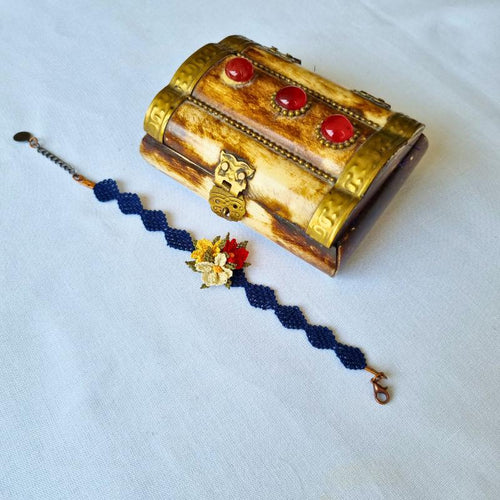 A handmade bracelet with three needle lace flowers in red, ivory, and yellow, with green leaves, on a dark blue band. Made with artificial silk thread using the Turkish Oya technique.