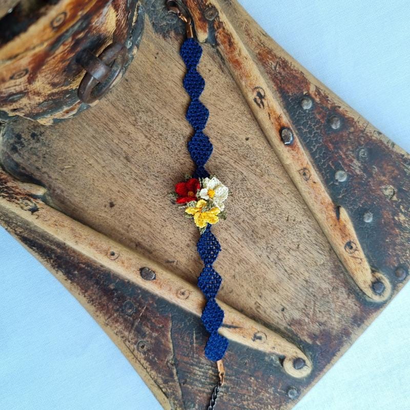 A handmade bracelet with three needle lace flowers in red, ivory, and yellow, with green leaves, on a dark blue band. Made with artificial silk thread using the Turkish Oya technique.